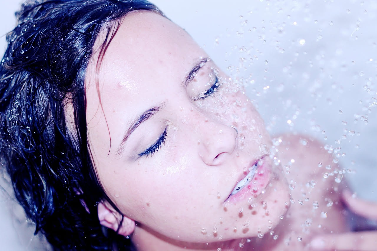 Capelli donna bagnati sotto il getto di doccia, con schiuma di shampoo, mani che massaggiano il cuoio capelluto in primo piano, specchio del bagno sullo sfondo
